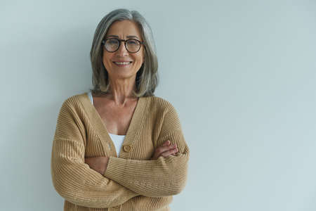 Elegant senior woman keeping arms crossed and smiling while standing against white wallの写真素材