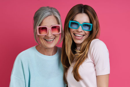 Happy mother and adult daughter in funky eyeglasses standing against pink background