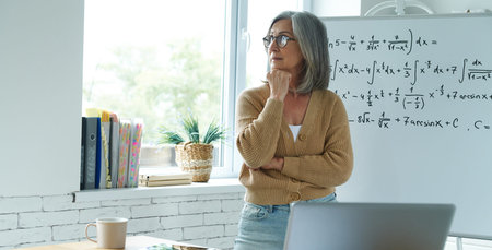 Thoughtful senior woman holding hand on chin while standing near the whiteboard at classroom