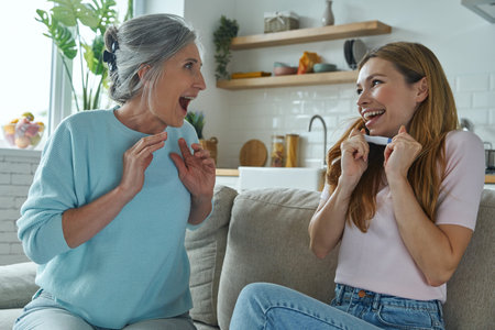 Happy young woman holding pregnancy test while sitting with her mother on the couch at home