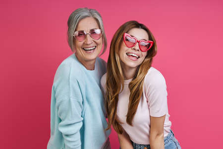 Happy mother and adult daughter wearing funky eyeglasses while standing together against pink backgroundの写真素材
