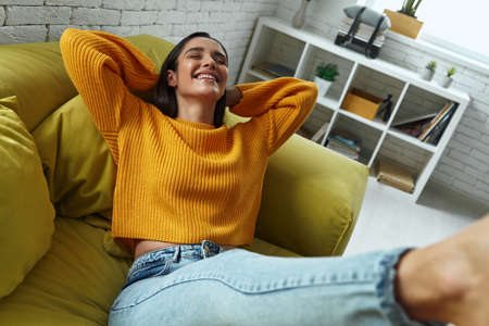 Joyful young woman holding hands behind head while sitting on the couch at homeの写真素材