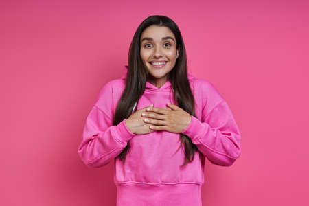 Surprised young woman in hooded shirt looking at camera and smiling against pink backgroundの写真素材