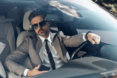 Confident mature man in formalwear holding smart phone while driving a car
