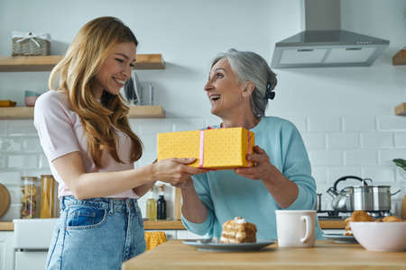 Surprised senior woman receiving a gift box from her daughter while sitting at the kitchen togetherの写真素材