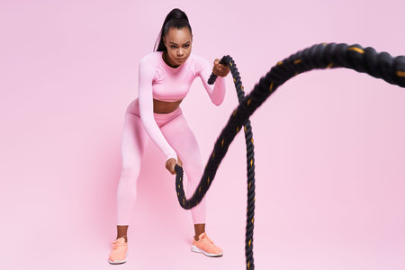 Confident young African woman exercising with battle ropes against pink backgroundの写真素材
