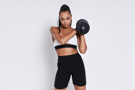 Confident young African woman holding dumbbell while standing against white background