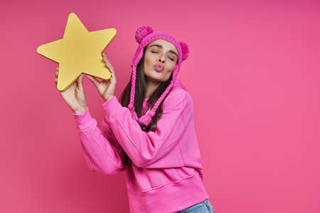 Playful young woman in funky hat holding paper star and puckering against pink backgroundの写真素材