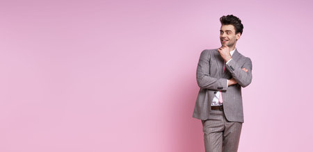 Cheerful young man in full suit holding hand on chin while standing against pink background