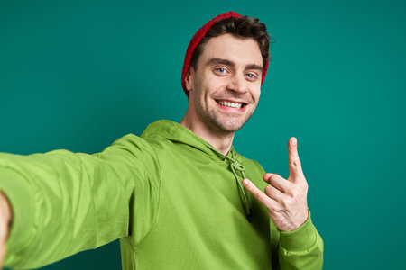 Cheerful young man making selfie and gesturing while standing against green background