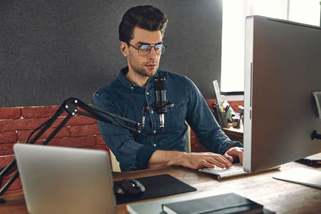 Confident young man using computer and microphone while recording podcast in studioの写真素材