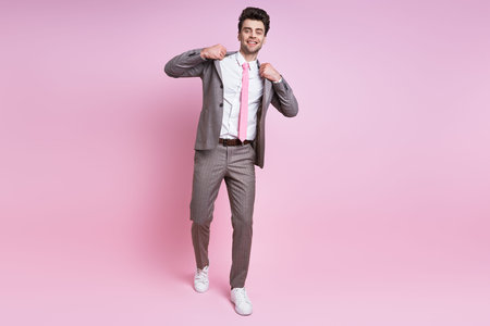 Cheerful young man in full suit adjusting his jacket while standing against pink background