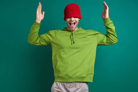 Happy young man pulling his hat on face and smiling while standing against green backgroundの写真素材