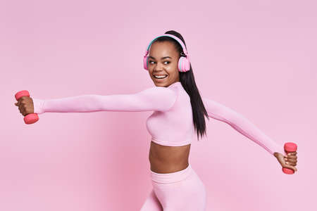 Cheerful African woman in headphones exercising with dumbbells against pink backgroundの写真素材
