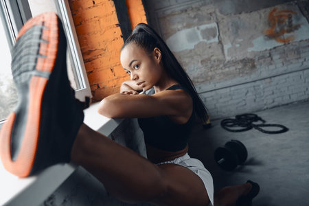 Confident woman in sports clothing using window sill while doing stretching exercises in gymの写真素材