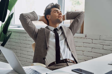 Relaxed young man in formalwear holding hands behind head while sitting in officeの写真素材