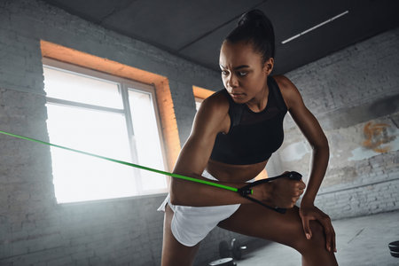 Beautiful young African woman exercising with elastic resistance band in gym