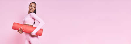 Happy African woman in sports clothing carrying exercise mat against pink backgroundの写真素材