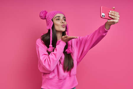 Playful young woman in funky hat making selfie and blowing a kiss against pink backgroundの写真素材