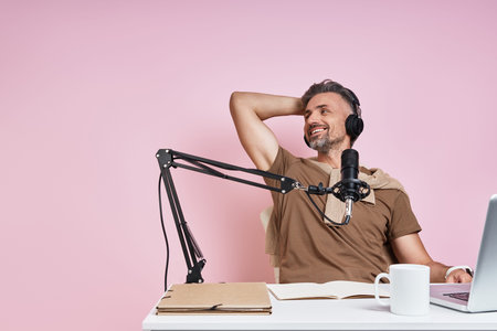 Relaxed man in headphones using microphone while recording podcast against pink background