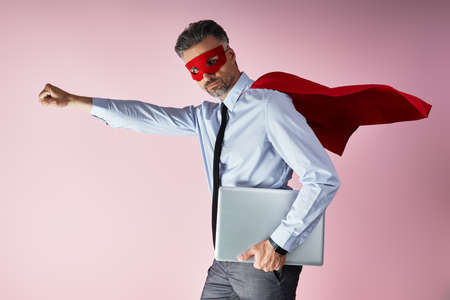 Confident man in shirt and tie wearing superhero cape and carrying laptop against pink backgroundの写真素材