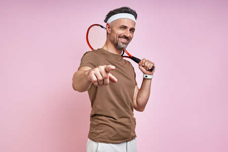 Happy mature man carrying tennis racket and pointing camera while standing against pink backgroundの写真素材