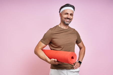 Happy mature man carrying exercise mat and smiling while standing against pink backgroundの写真素材