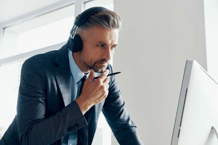 Thoughtful mature businessman in headphones looking at the computer while standing in officeの写真素材