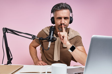 Serious mature man in headphones holding finger on lips while recording podcast against pink...