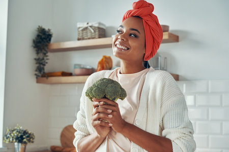 Beautiful young African woman holding broccoli and smiling while standing at the domestic kitchenの写真素材