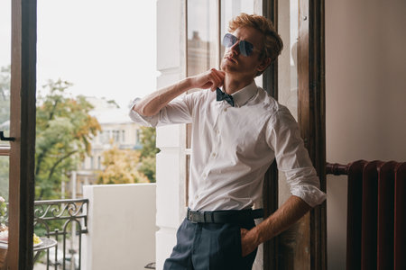 Handsome young man in in shirt and bowtie adjusting his shirt while leaning at the balcony doorの写真素材