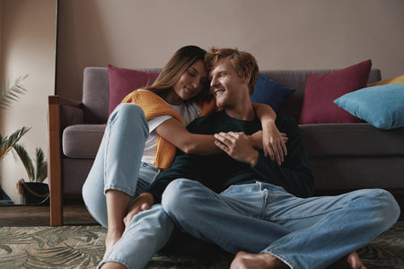 Beautiful young couple embracing and smiling while relaxing at home togetherの写真素材