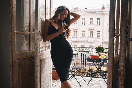 Gorgeous woman in cocktail dress holding flute with champagne while leaning at the balcony doorの写真素材