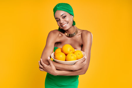 Attractive African woman in traditional headwear holding oranges and smiling against yellow backgroundの写真素材