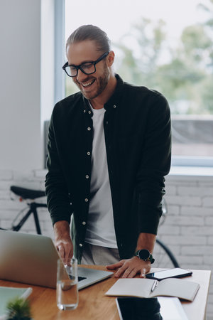Cheerful young man using laptop while standing near his working place in officeの写真素材