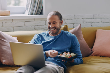 Cheerful young man eating lunch and looking at laptop while sitting on the couch at homeの写真素材
