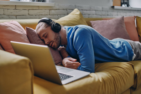Young man in headphones sleeping near his laptop on the couchの写真素材