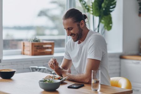Cheerful young man enjoying lunch while sitting at the kitchen counter at homeの写真素材