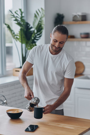 Handsome young man pouring coffee to the cup and smiling while standing at the kitchenの写真素材