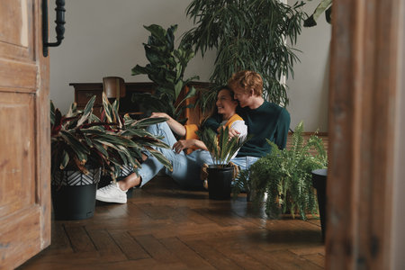 Beautiful young couple watering houseplants while sitting on the floor at homeの写真素材