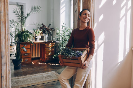 Happy young woman carrying wooden crate with plants while leaning at the doorway at homeの写真素材