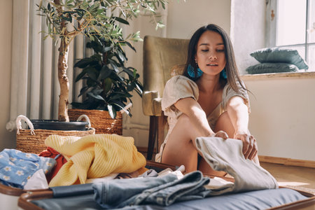 Beautiful young woman packing clothes to suitcase while sitting on the floor at homeの写真素材