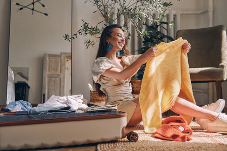 Attractive young woman packing clothes to suitcase and smiling while sitting on the floor at homeの写真素材