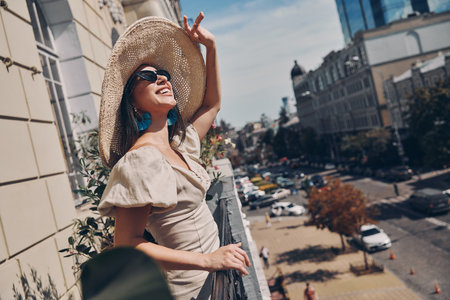 Relaxed young woman adjusting her elegant hat and smiling while standing on the balconyの写真素材