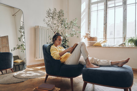 Attractive young woman reading a book while relaxing in a comfortable chair at homeの写真素材