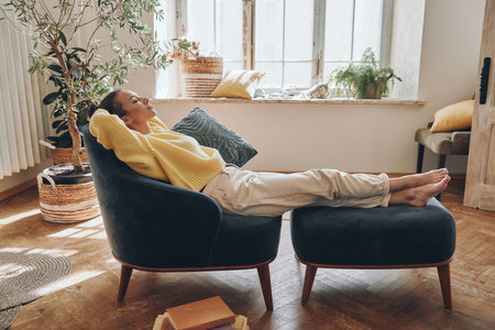 Beautiful young woman holding hands behind head while relaxing in comfortable chair at homeの写真素材