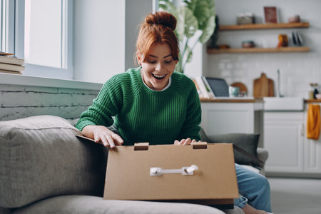 Happy young woman unpacking box while sitting on the couch at homeの写真素材