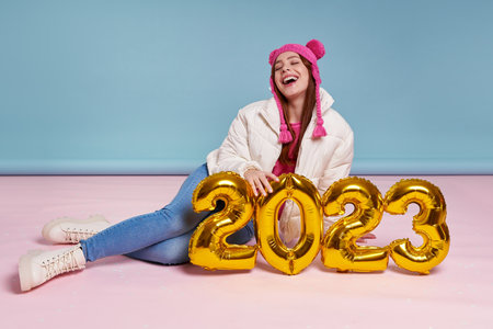 Cheerful young woman in winter coat holding gold colored numbers while sitting against blue backgroundの写真素材