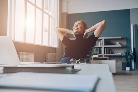 Relaxed businesswoman holding hands behind head and smiling while sitting at the office deskの写真素材