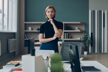 Serious businesswoman looking at camera while standing near her working place in officeの写真素材